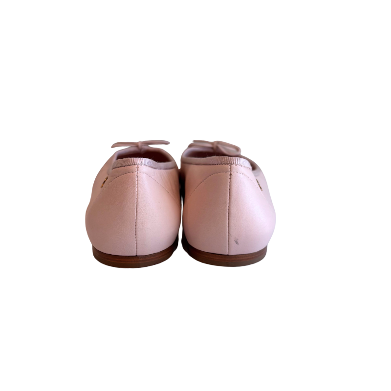 A pair of light pink leather ballet flats shown from the back, featuring a small bow at the top of each shoe and a brown sole, set against a plain white background.