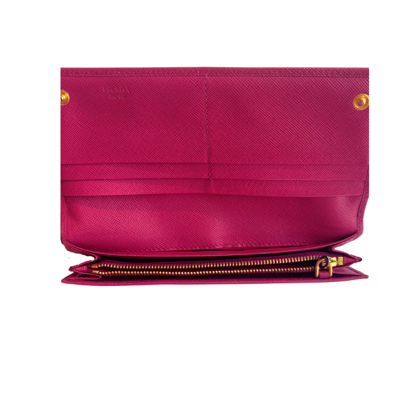 A close-up of a magenta pink textured leather wallet or clutch shown open, featuring gold-tone hardware, a zippered compartment, and a visible 'PRADA' logo on the upper left.