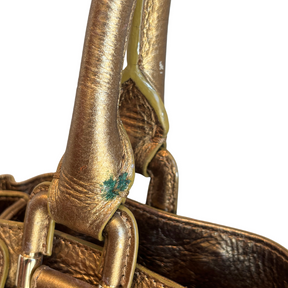 Close-up detail of a metallic gold leather handbag handle with a small green painted mark, set against a plain white background.