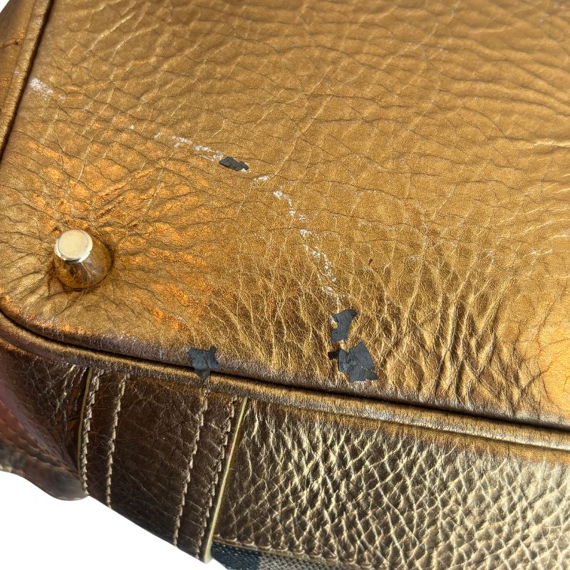 Close-up of a gold-toned textured leather handbag corner showing a brass rivet and visible scuffs and wear on the leather surface.