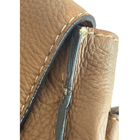 Close-up of a tan leather bag corner showing textured leather, cream stitching, and a dark strap attachment with visible wear and tear.